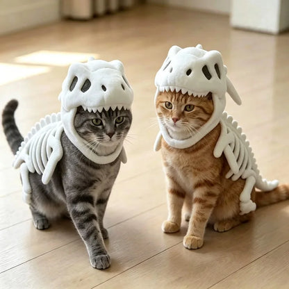 Two cats wearing plush dinosaur skeleton costumes, sitting on a light wood floor in a bright indoor setting.