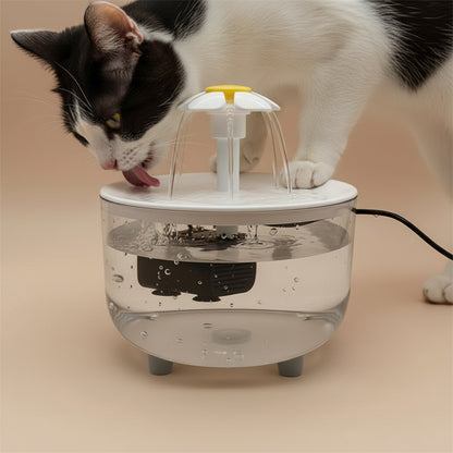 Cat drinking from A small tabletop water fountain with a clear cylindrical basin, white top and base, a central white spout with a yellow ring, and a black pump submerged in the water. A black power cord extends to the right.