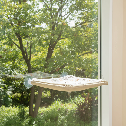 A beige fabric cat hammock suspended by cords from a clear suction cup window perch, with a bright window and city view in the background.